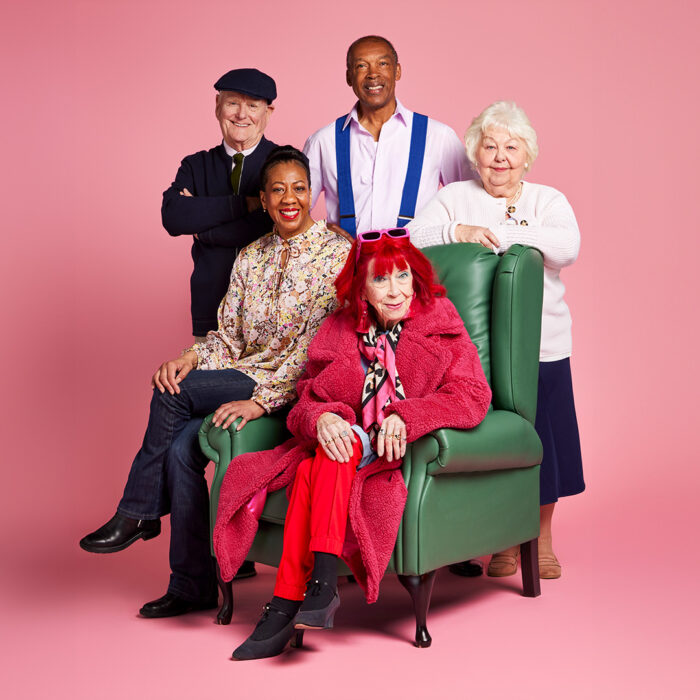 Photo of 5 elderly men and women stood around a green arm chair smiling, with a pink backdrop behind them.