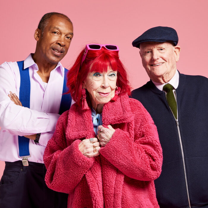 Photo of 2 elderly men and 1 woman stood smiling, with a pink backdrop behind them.